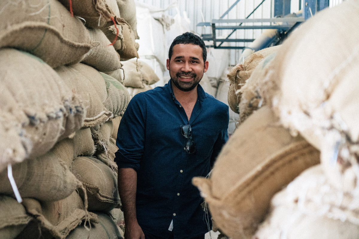 A smiling man in a dark shirt standing between tall stacks of coffee sacks